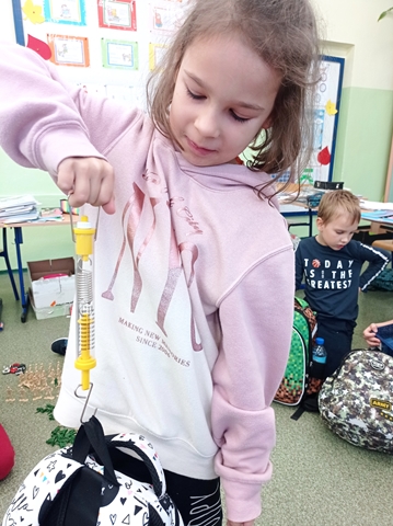 Zdjęcie przedstawia dziewczynkę w biało-różowej bluzie ważącą swój plecak specjalnym urządzeniem.