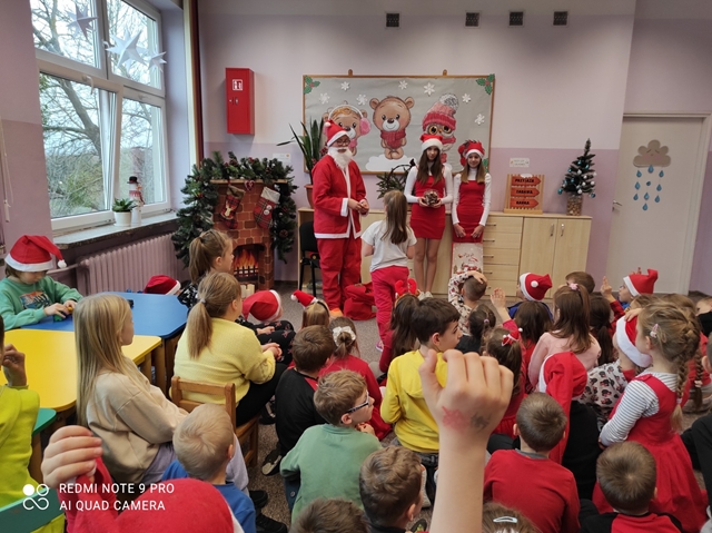 Zdjęcie przedstawia grupę dzieci uczestniczących w spotkaniu ze świętym Mikołajem w świetlicy szkolnej. Uczniowie mają na sobie ubrania w kolorze czerwonym i mikołajowe czapki.