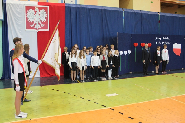 Uczniowie ubrani w galowe stroje stoją na baczność. Wszystkie dzieci śpiewają hymn. 