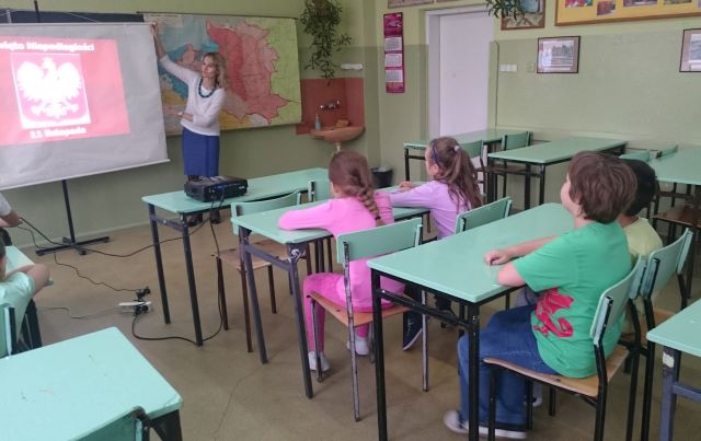 Zdjęcie przedstawia uczniów siedzących w szkolnych ławkach. Na środku sali nauczycielka wskazuje na tablicę z godłem Polski. W tle ukazana jest mapa Polski. 