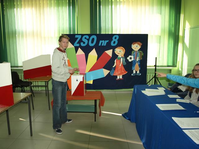Zdjęcie przedstawia ucznia wrzucającego swój głos do urny wyborczej. 