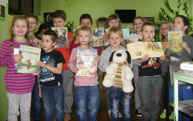 Zdjęcie przedstawia uczniów w bibliotece szkolnej. Dzieci prezentują książki nawiązujące do Dnia Pluszowego Misia. Jeden z chłopców trzyma dużą maskotkę.