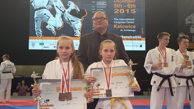 Zdjęcie przedstawia dwie uczennice występujące w strojach karate. Dziewczynki trzymają dyplomy i statuetki, a na ich szyjach widnieją zdobyte medale. W tle widać innych uczestników zawodów.