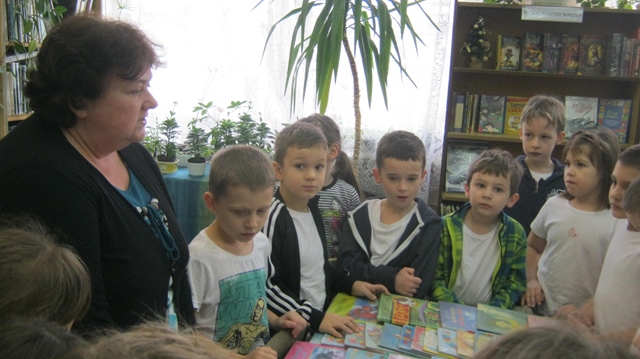 Zdjęcie przedstawia dzieci  z panią bibliotekarką w bibliotece szkolnej stojące dookoła stołu z książkami. 