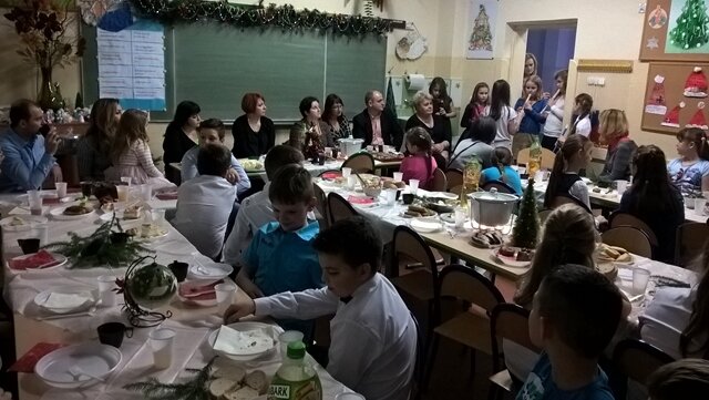 Uczniowie i rodzice w sali lekcyjnej podczas wigilii klasowej. Wszyscy siedzą przy zastawionych stołach potrawami wigilijnymi.