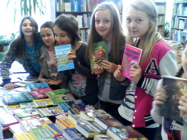 Zdjęcie kilku uczennic w bibliotece szkolnej prezentujących książki, które można wypożyczyć. Na stole przed dziewczynkami znajduje się wystawka tych książek