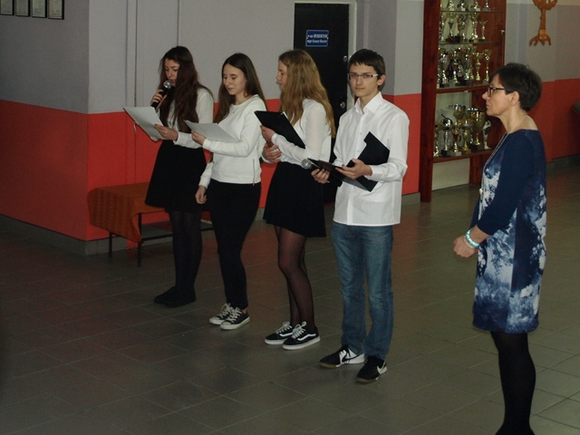 Zdjęcie przedstawia uczniów prowadzących apel w holu głównym szkoły. 