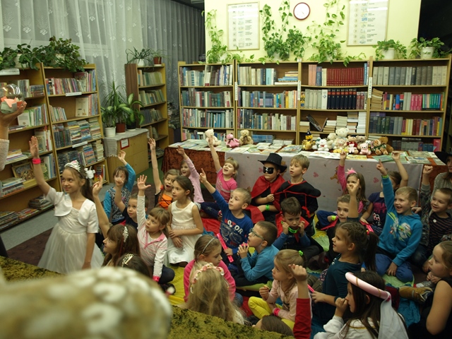 Zdjęcie przedstawiające uczniów aktywnie uczestniczących w zajęciach w bibliotece szkolnej. Niektóre dzieci są przebrani za postacie z bajek. W tle regały z książkami i stół, na którym znajdują się maskotki i książki.