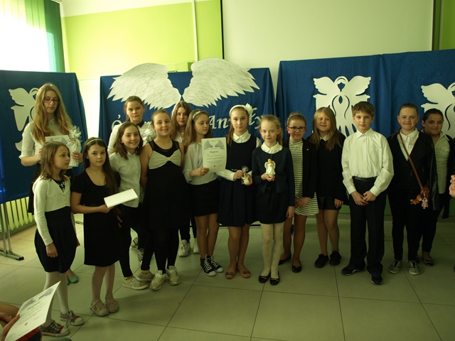 Zdjęcie grupy uczniów w strojach galowych. Niektórzy uczniowie trzymają w dłoniach dyplomy lub figurki aniołów. W tle tablice udekorowane anielskimi skrzydłami.