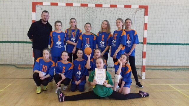 Zdjęcie przedstawia uczennice w strojach sportowych z nauczycielem wychowania fizycznego. Wszyscy stoją w sali gimnastycznej na tle bramki.