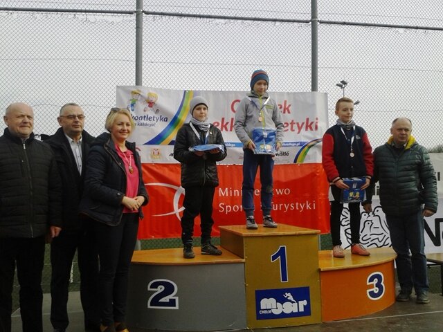 Zdjęcie przedstawia zdobywcę pierwszego miejsca w biegach przełajowych. Zwycięzca stoi na podium i dumnie prezentuje złoty medal oraz nagrodę rzeczową.
