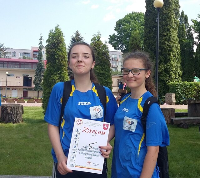 Zdjęcie przedstawia dwie zawodniczki w strojach sportowych. Dziewczęta prezentują dyplom zdobyty w zawodach.