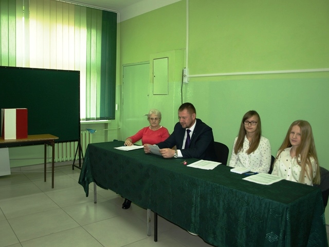 W sali lekcyjnej komisja wyborcza, w skład której wchodzą dwie osoby dorosłe i dwie uczennice reprezentujące społeczność szkolną. W tle biało – czerwona urna wyborcza.