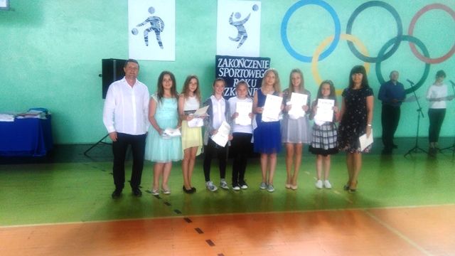 Zdjęcie przedstawia grupę uczennic wraz z nauczycielami. Dziewczęta prezentują dyplomy uznania w sali gimnastycznej. W tle tablica z napisem “Zakończenie sportowego roku szkolnego”.