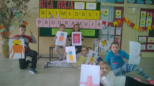 Grupa uczniów prezentująca swoje prace. W tle dekoracyjna tablica z napisem „Mega Misja – najmłodsi programują”.