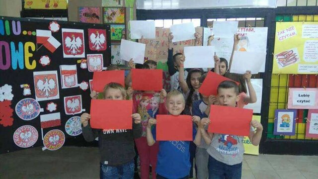 Uczniowie trzymają w rękach białe i czerwone kartki, tworząc flagę państwową. W tle tablica udekorowana godłami Polski wykonanymi przez uczniów.