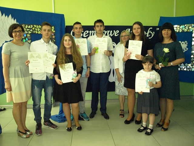 Zdjęcie grupy uczniów z nauczycielami i panią dyrektor. Dzieci w strojach galowych trzymają dyplomy. W tle tablica dekoracyjna.