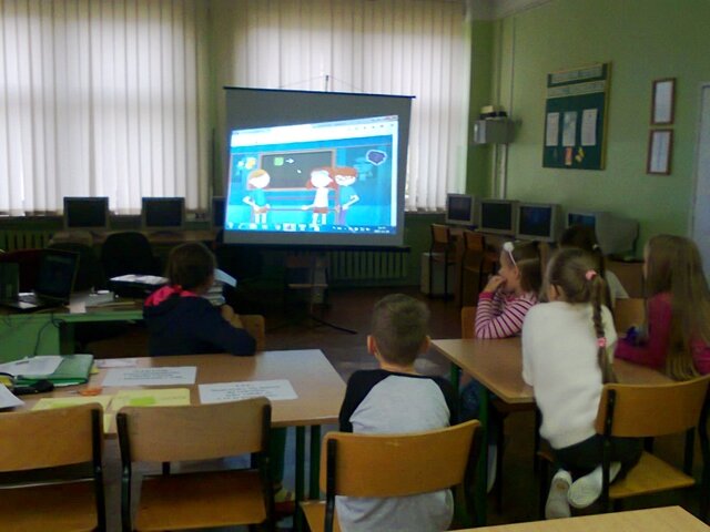 Dzieci siedzą w ławkach i oglądają film wyświetlany na dużym ekranie.