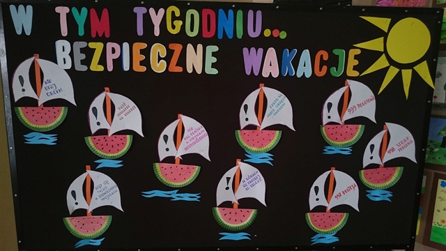 Dekoracyjna tablica z napisem „W tym tygodniu... bezpieczne wakacje”. Do tablicy zostały przypięte słońce i łódki, których kadłub stanowią połówki arbuza, a na żaglach rady dotyczące bezpieczeństwa.