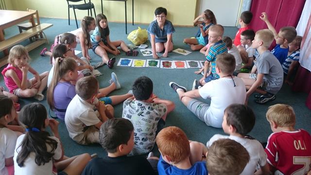 Zdjęcie przedstawia uczniów i nauczycielkę  podczas zajęć  na temat segregacji odpadów. Na dywanie rozłożone rysunki pojemników na śmieci w różnych kolorach. 