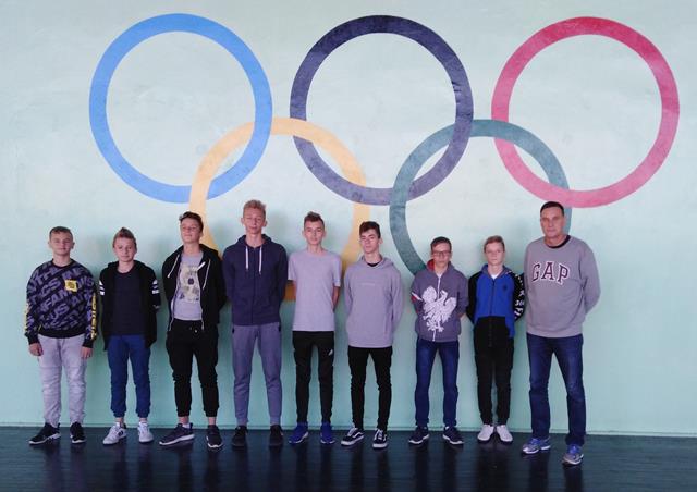 Zdjęcie przedstawia grupę chłopców oraz nauczyciela stojących na tle ściany z flagą olimpijską. 
