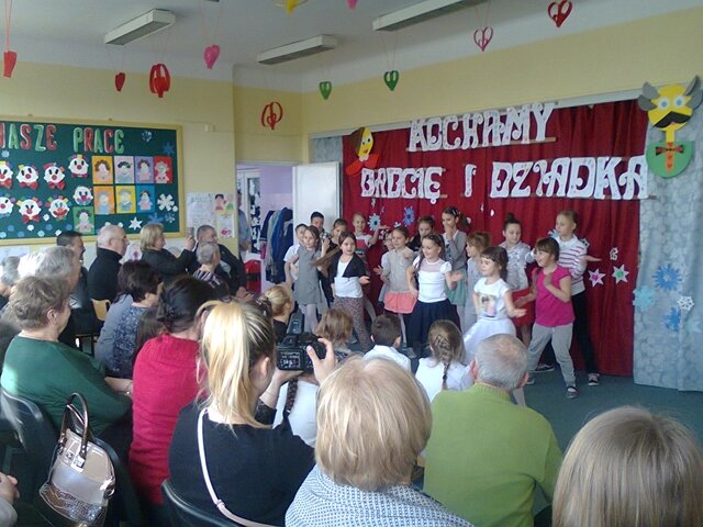 Zdjęcie przedstawiające uczniów i zaproszonych gości podczas obchodów Dnia Babci i Dziadka w świetlicy szkolnej. Uczniowie stoją na tle dekoracji, babcie i dziadkowie z zainteresowaniem oglądają występ.