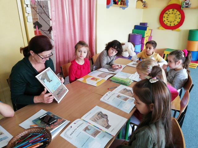 Zdjęcie przedstawiające uczniów i nauczycielkę podczas zajęć w świetlicy szkolnej. Dzieci siedzą przy stolikach, przed nimi otwarte zeszyty ćwiczeń.