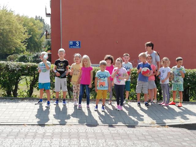 Zdjęcie przedstawia uczniów z nauczycielką stojących przed przejściem dla pieszych podczas zajęć na temat bezpieczeństwa.