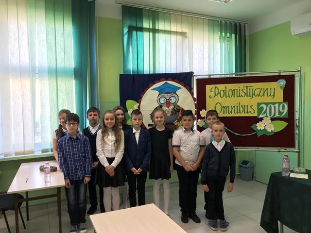 Zdjęcie przedstawia uczestników konkursu, stojących przed tablicami z napisem „Polonistyczny Omnibus 2019”.