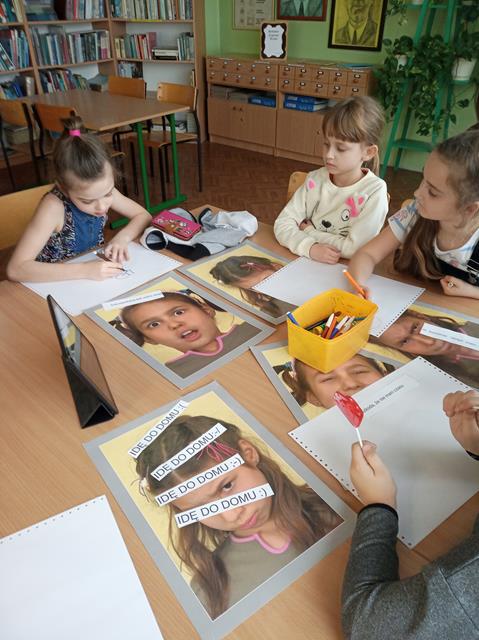 Czworo dzieci siedzi przy stoliku, na którym leżą kartony, zdjęcia dziewczynek wyrażających mimiką twarzy różne emocje oraz napisy „Idę do domu” zakończone emotkami, stoi pudełko z pisakami. Dwie uczennice rysują.