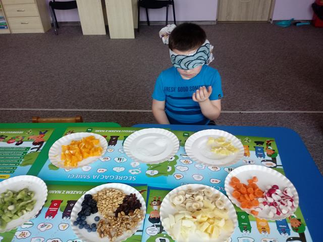 Chłopiec z zawiązanymi oczami siedzi przy stoliku, na którym stoją jednorazowe talerze z różnymi  pokrojonymi owocami i warzywami.