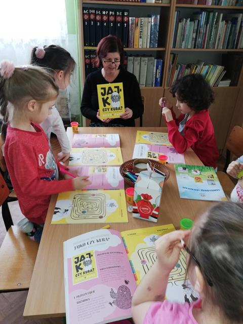 Pani bibliotekarka wraz z dziećmi, które rozwiązują „Łamigłówki Literackie” do książki „Jajko czy kura”.