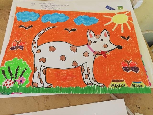 Praca plastyczna wykonana przez ucznia. Na rysunku biały piesek w brązowe łatki przy dwóch miseczkach. Tło jest pomarańczowe z motylkami, ptakami, niebieskimi chmurami i słońcem.