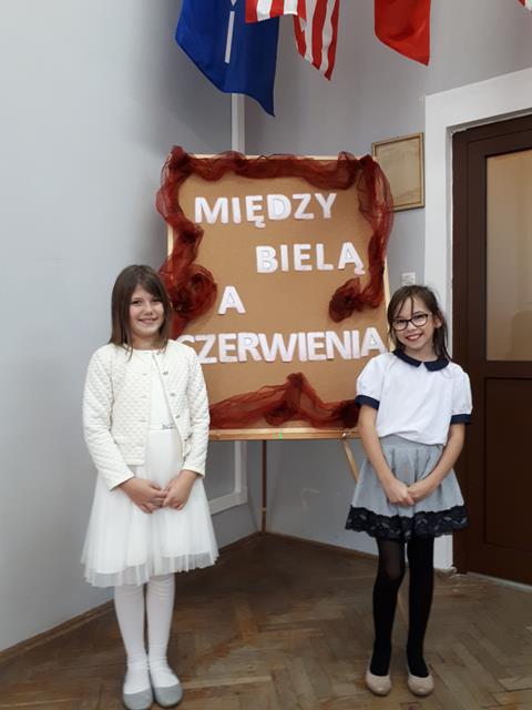 Zdjęcie przedstawia dwie uczennice ubrane na galowo. Dziewczynki stoją na tle tablicy z napisem „Między bielą a czerwienią”.