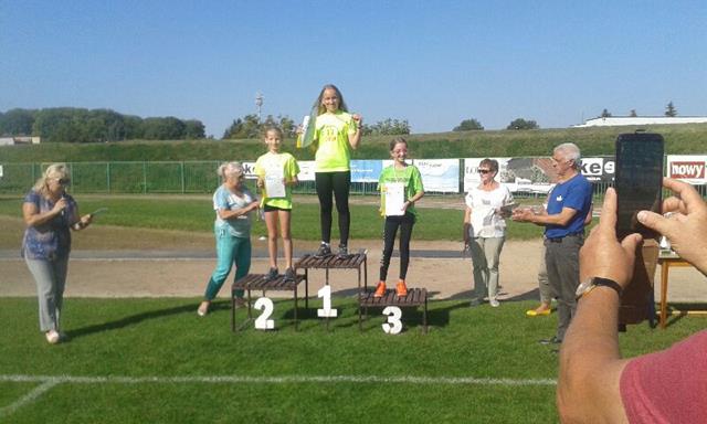 Zdjęcie wykonane na Stadionie Miejskim w Chełmie przedstawia usytuowane na podium finalistki zawodów z medalmi i dyplomami. Na najwyższym stopniu stoi zawodniczka z Ósemki. W tle widoczni są organizatorzy zawodów.