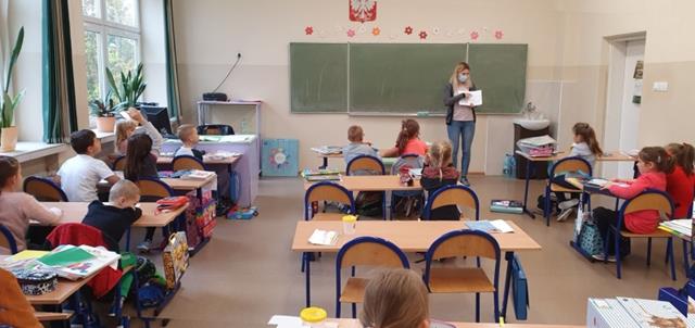 Zdjęcie klasy. Uczniowie siedzą w ławkach, a przy tablicy na środku klasy stoi nauczyciel i pokazuje coś w książce.
