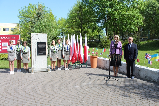 Pani Dyrektor w obecności zaproszonego gościa przemawia w czasie obchodów 78 rocznicy wyzwolenia Stalagu 319. Sześć harcerek stoi obok pomnika i flag państwowych.