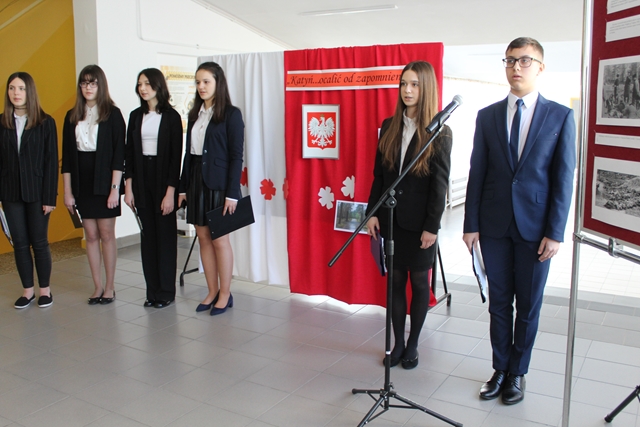 Uczniowie w strojach galowych stoją na tle biało-czerwonej tablicy opatrzonej hasłem „Katyń – ocalić od zapomnienia...”
