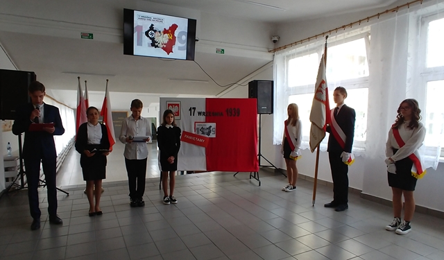 Zdjęcie przedstawia uczniów podczas apelu w holu szkoły. Po prawej stronie stoi poczet sztandarowy, po lewej  uczniowie prowadzący apel. W tle okolicznościowa dekoracja.