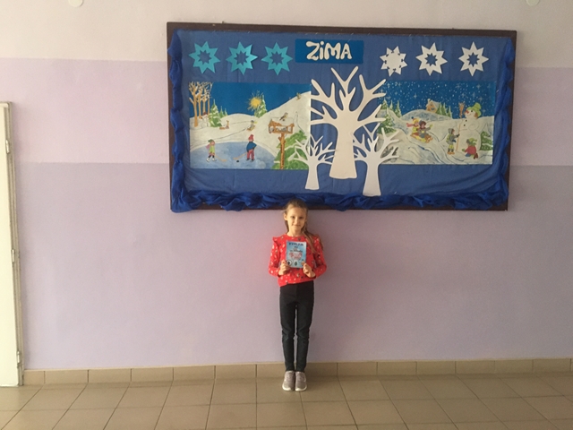 Zdjęcie przedstawia uczennicę, która prezentuje dyplom z konkursu. W tle tablica z zimową dekoracją.