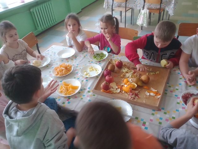 Uczniowie przygotowują owocową sałatkę. W talerzykach na stole leżą różnokolorowe owoce. Dzieci obierają, kroją oraz układają wszystkie składniki.