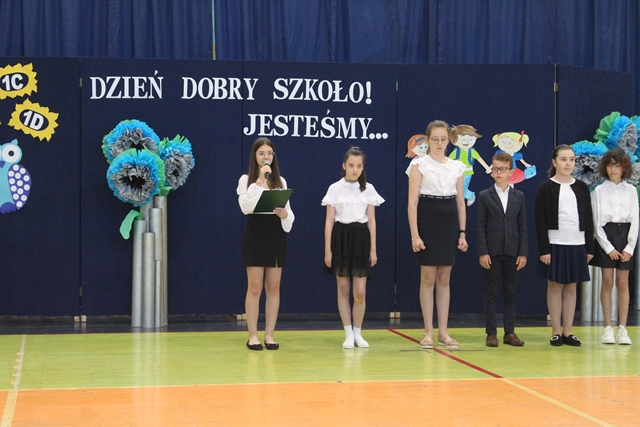 Zdjęcie przedstawia kilkoro uczniów podczas uroczystości rozpoczęcia roku szkolnego. W tle dekoracja z napisem : "Dzień dobry, szkoło! Jesteśmy…’’.