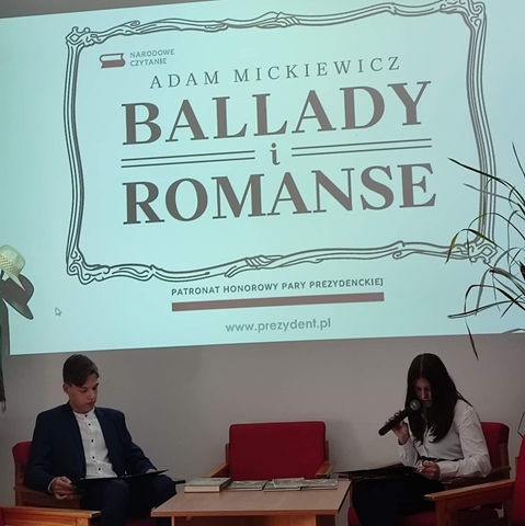 Zdjęcie przedstawia dwoje uczniów siedzących przy stoliku podczas Narodowego Czytania. Uczennica czyta książkę, w dłoni trzyma mikrofon. Nad nimi wyświetlony napis ,, Adam Mickiewicz - Ballady i romanse’’.