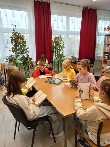 Zdjęcie prezentujące uczniów w siedzących przy stoliku i czytających książki w bibliotece szkolnej.