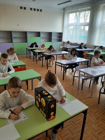 Zdjęcie przedstawia uczniów podczas konkursu. Dzieci siedzą w ławkach i uzupełniają arkusze konkursowe.