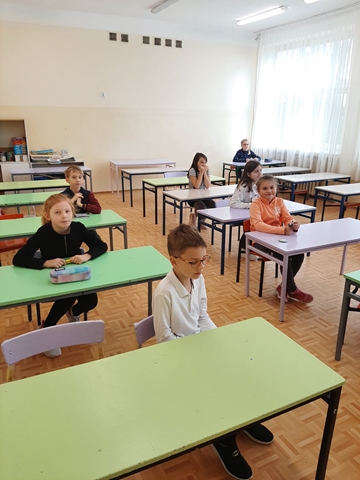 Zdjęcie przedstawia uczniów klas młodszych podczas konkursu. Dzieci siedzą w ławkach i czekają na rozdanie arkuszy. 