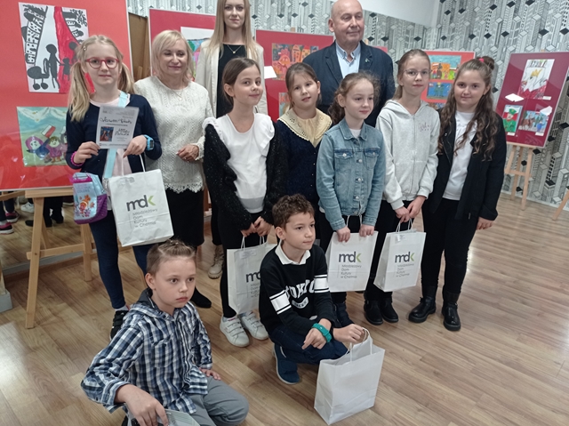 Zdjęcie przedstawia uczniów klas 1-3 biorących udział w konkursie plastycznym „Młody Patriota”. Dzieci trzymają papierowe torebki z nagrodami i wraz z nauczycielami pozują do zdjęcia.