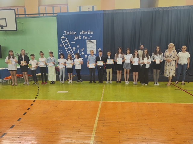 Zdjęcie przedstawia  grupę uczniów w sali gimnastycznej  podczas podsumowania konkursów. Laureaci prezentują otrzymane dyplomy i nagrody.