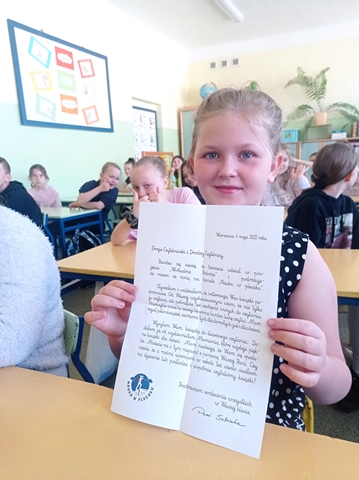 Grupa dzieci siedzi w sali lekcyjnej. Jedna dziewczynka trzyma przed sobą list.