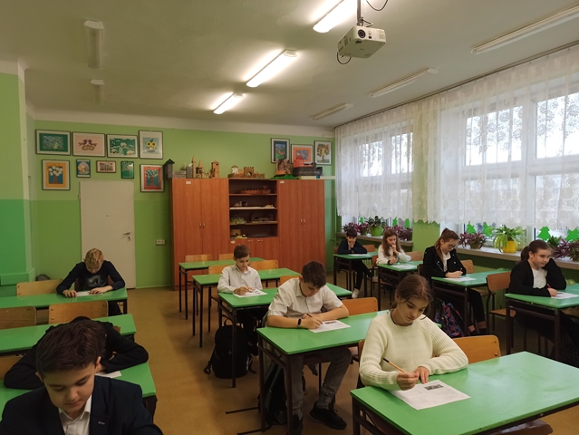 Zdjęcie przedstawia uczniów klas starszych podczas konkursu. Uczniowie siedzą w ławkach i uzupełniają konkursowe arkusze.
